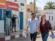 Traum vom Auswandern nach Djerba: Glückliches Paar bei der Erkundung der Insel, begleitet von freundlichen Einheimischen, vor typischer weiß-blauer Architektur, für alle, die einen Umzug nach Djerba planen.
